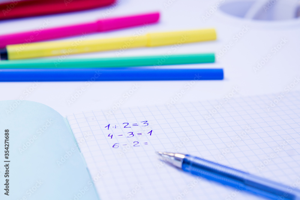 On the white table of the student, arithmetic examples are written in an open notebook in a cage. Also on the table are colored pencils and markers. On the white table of the student, arithmetic examples are written in an open notebook in a cage. Also on the table are colored pencils and markers.