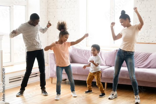 Full length view active African ethnicity couple and their kids daughter and son dancing standing in modern living room, homeowners enjoy free time together listen music spend funny activities at home