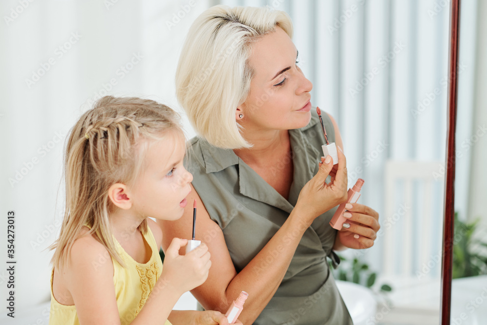 Mother applying liquid lipstick in front of mirror and her daughter repeating after her Mother applying liquid lipstick in front of mirror and her daughter repeating after her