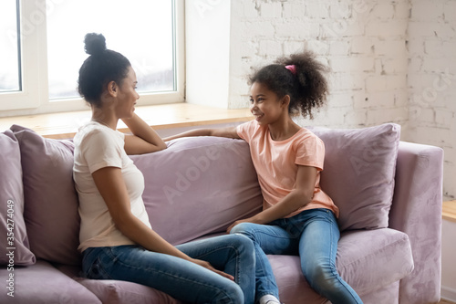 African mother and pre-teen daughter sitting together on sofa having honest pleasant conversation. Concept of trusting relationship between kid and parent, gossips secrets sharing, heart-to-heart talk