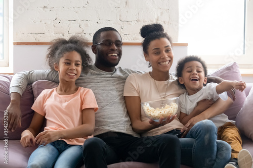 Full African ethnicity family, spouses with daughter and son spend weekend free time together at home sitting on couch in living room eating popcorn watching TV show movie or cartoons have fun concept