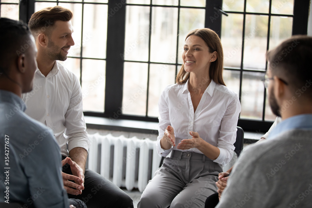 Fototapeta premium Confident smiling woman coach speaking at meeting, happy diverse people sitting in circle at group therapy session with psychologist, mentor training staff, team building activity in office