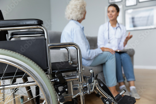 Physical therapist woman in white coat talk to elderly woman disabled patient client seated on sofa in living room, close up focus on wheelchair. Concept of handicapped person, caregiving and homecare