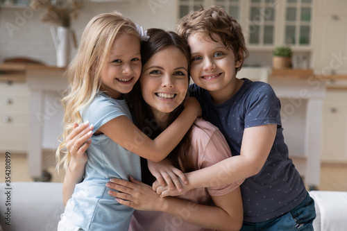 Portrait of happy young mother hug cuddle with smiling little children relax on weekend at home, smiling overjoyed small kids embrace mom or nanny show love and gratitude, family bonding concept