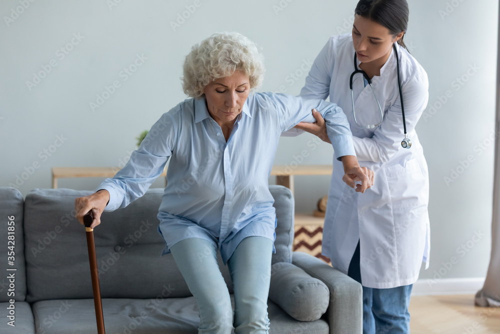 Obraz premium Caring young nurse helping to elderly disabled with walking cane woman get off the couch, age related disease, after illness or operation. Caregiving, provides physical support to old patient concept