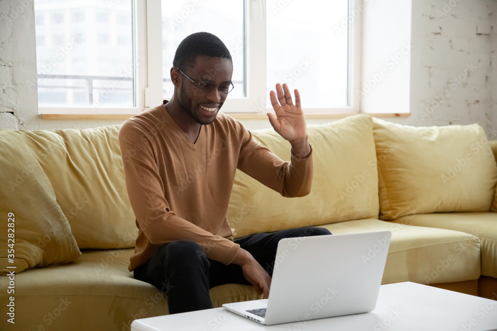 Φωτογραφία African millennial guy sit on couch makes videocall looks at pc screen wave hand feels happy glad to see greeting friend