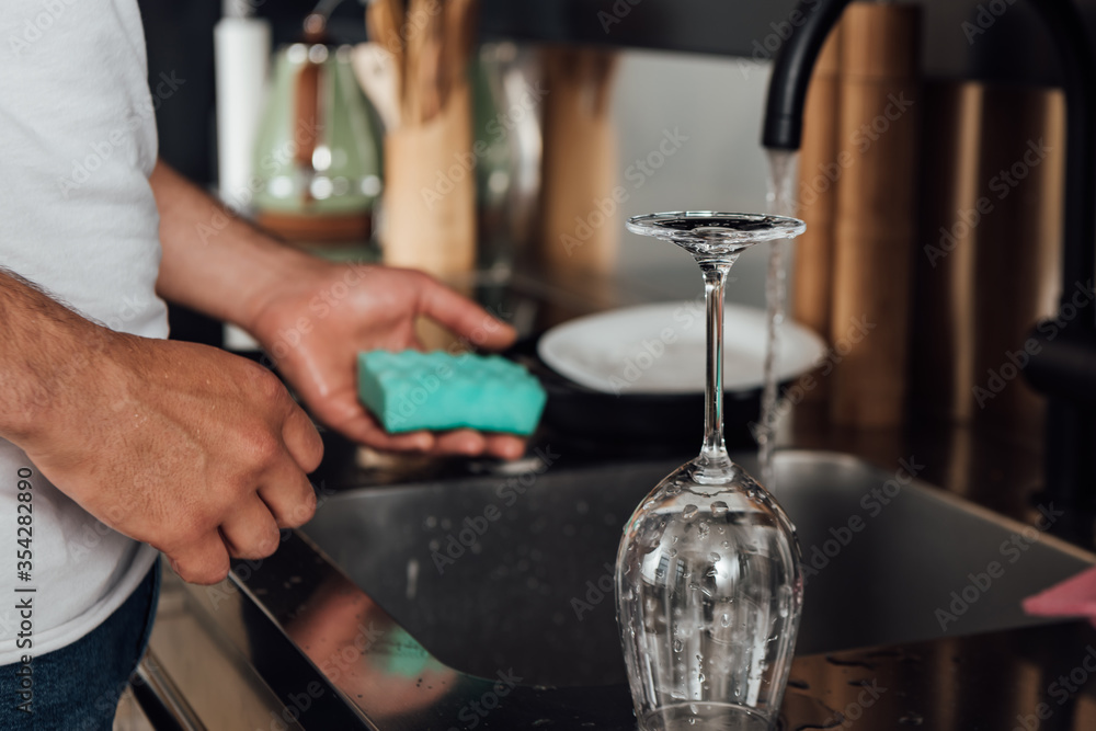 Cropped view of man holding sponge near wet wine glass and kitchen sink