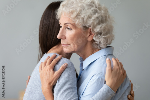 Close up young relative granddaughter visit hug elderly grandmother with distant a look supports her in hard period of life caused by senile disease or tragedy. Care, help, share pain, be near concept