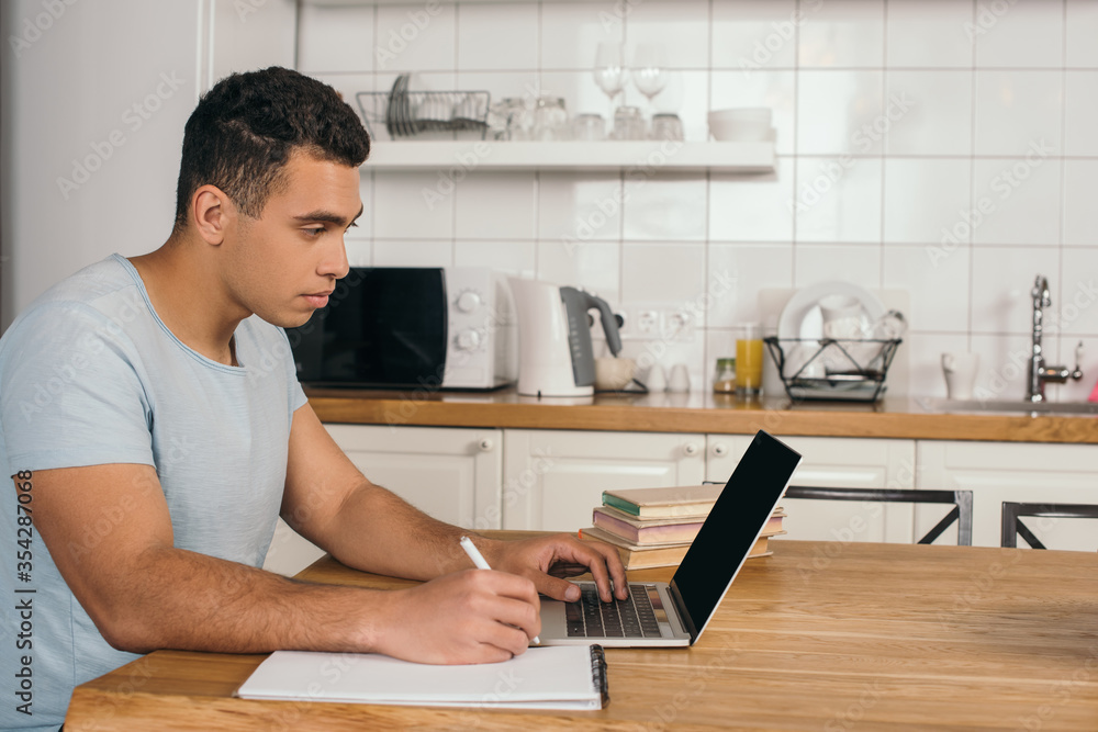 handsome mixed race man holding pen near notebook and using laptop with blank screen handsome mixed race man holding pen near notebook and using laptop with blank screen