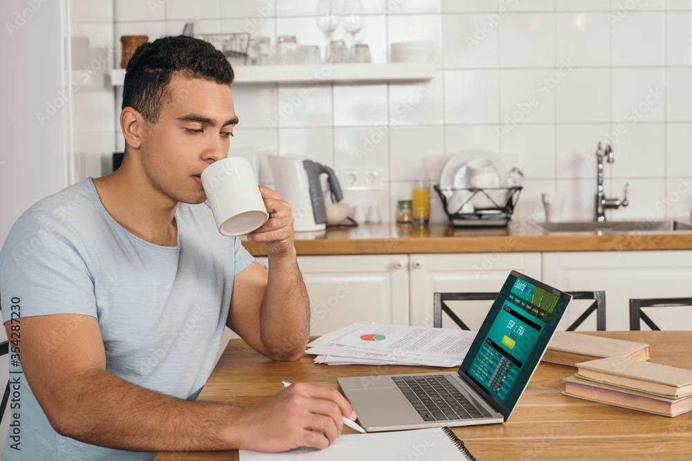handsome mixed race man holding pen and drinking coffee near laptop with sports bet website handsome mixed race man holding pen and drinking coffee near laptop with sports bet website