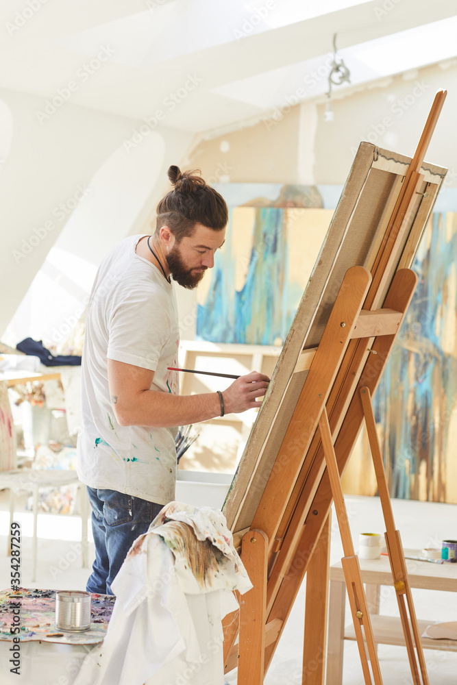 Vertical portrait of contemporary male artist painting picture on easel while standing in attic art studio lit by sunlight Vertical portrait of contemporary male artist painting picture on easel while standing in attic art studio lit by sunlight