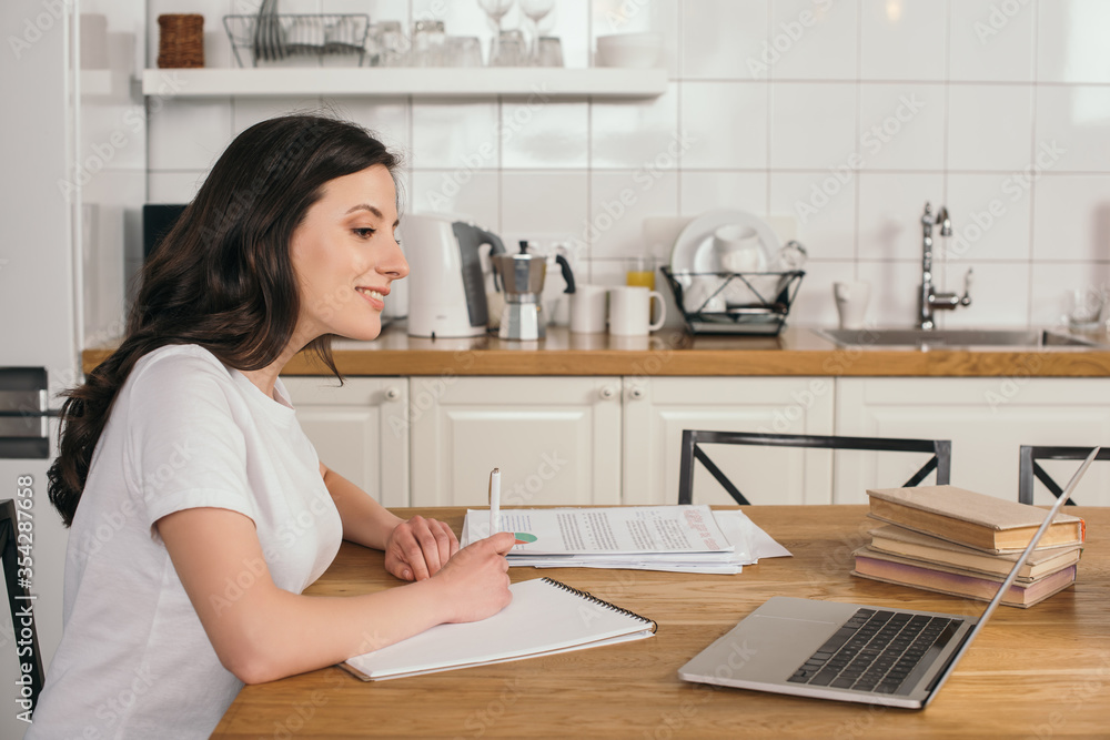 cheerful freelancer looking at laptop and writing in notebook, online study concept cheerful freelancer looking at laptop and writing in notebook, online study concept