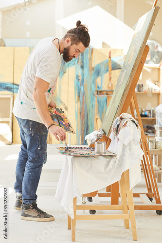 Full length portrait of contemporary bearded artist painting picture on easel while working in spacious art studio lit by sunlight