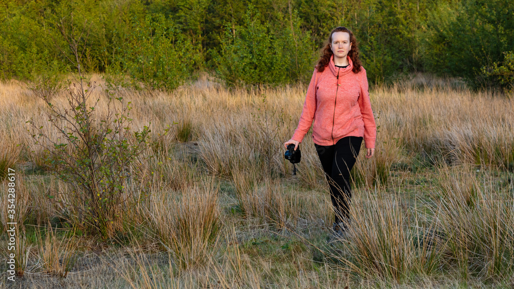 Young woman walking through a meadow carrying a camera, in late evening light.