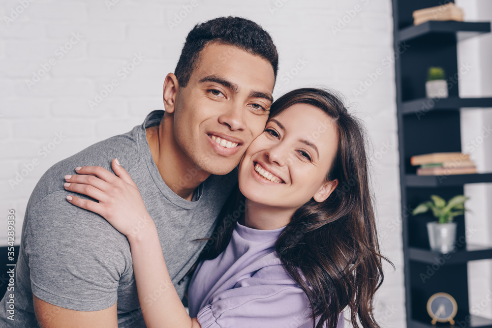 Obraz premium cheerful woman hugging handsome bi-racial man in bedroom