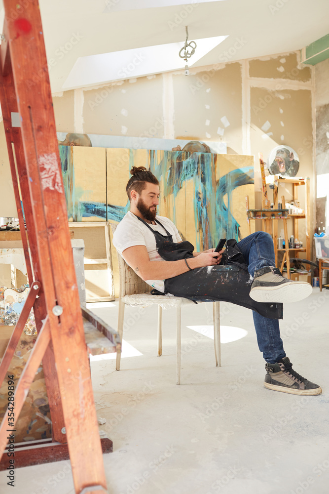 Vertical full length portrait of contemporary bearded artist using smartphone while sitting on chair in art studio with abstract paintings in background, copy space