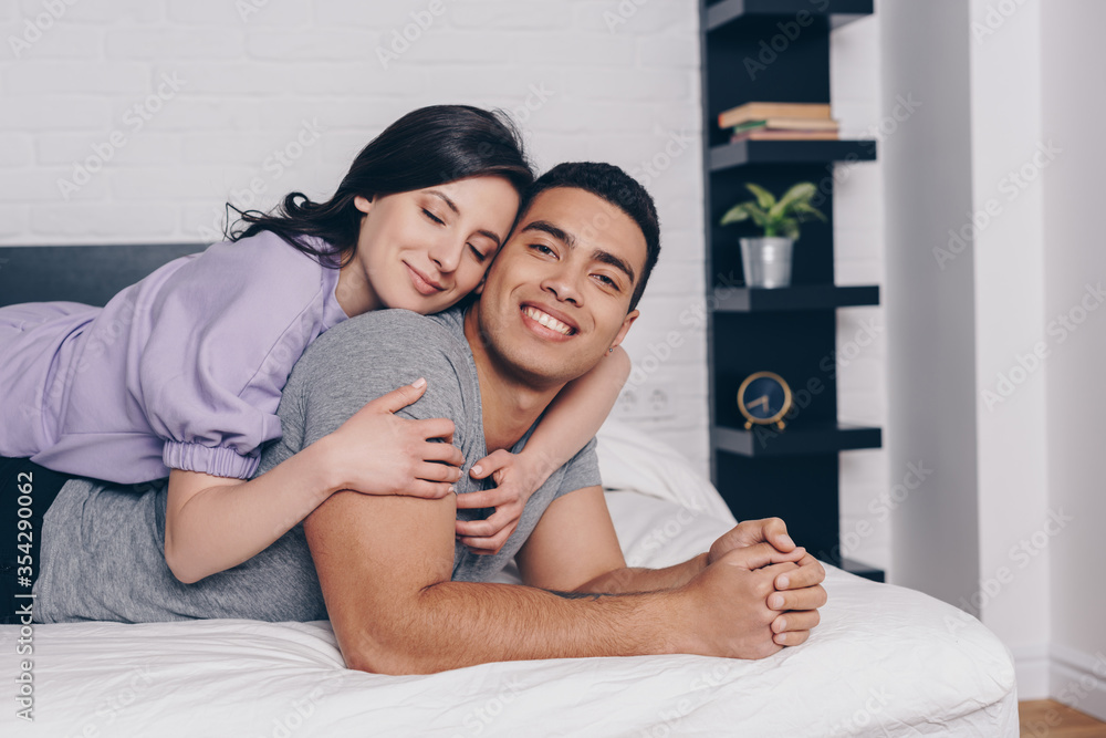 cheerful woman with closed eyes hugging handsome bi-racial man while lying on bed - obrazy, fototapety, plakaty 