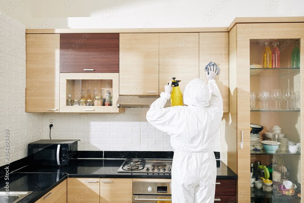 Worker in white protective suit wiping doors of kitchen cabinets with disinfecting spray Worker in white protective suit wiping doors of kitchen cabinets with disinfecting spray