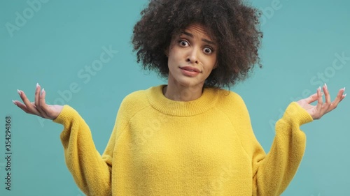 A confused young african american woman with curly hair is raising her hands isolated over blue wall background in studio