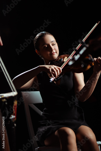 happy female musician with closed eyes playing on violin on dark stage