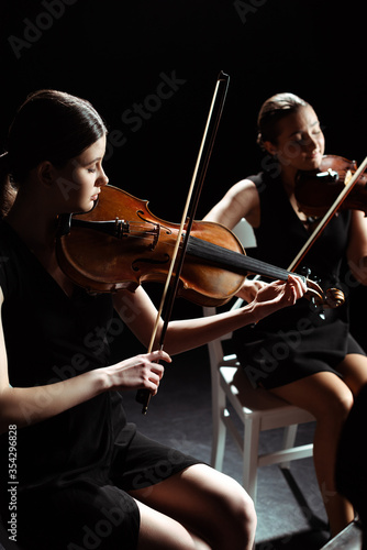 attractive professional female musicians playing classical music on violins on dark stage