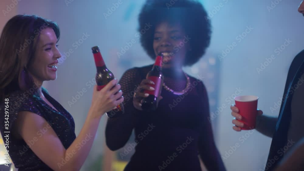 Handheld slowmo of happy young women holding beer bottles and laughing while dancing with friends at house party Handheld slowmo of happy young women holding beer bottles and laughing while dancing with friends at house party