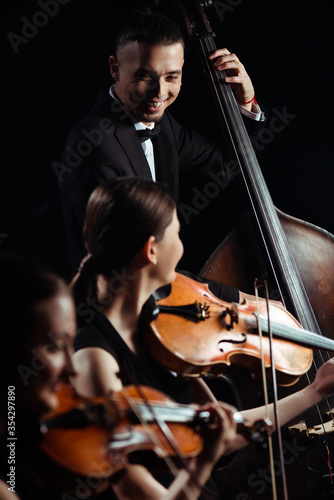trio of smiling musicians playing on double bass and violins isolated on black