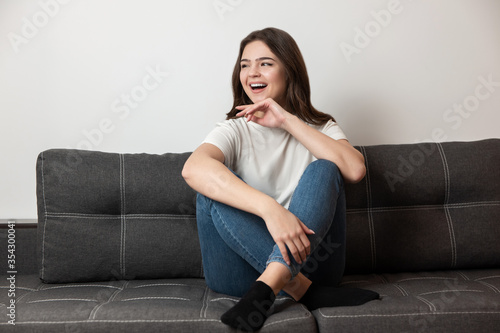 young beautiful smiling brunette woman sitting on the sofa in her appartment looking happy, feels cozy at home
