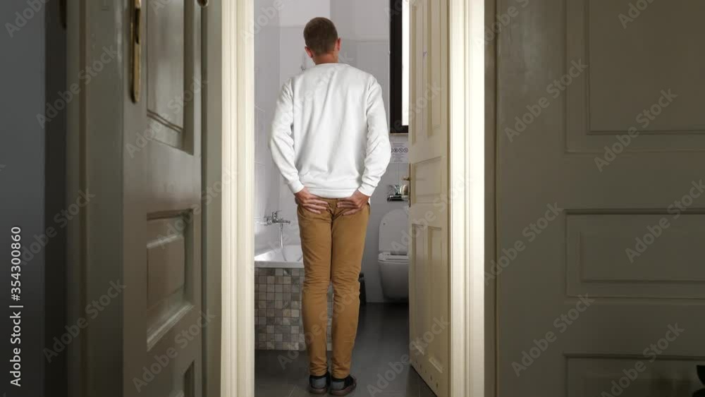 Young man enters bathroom and feels sudden pain in his butt, holds and rub buttocks Young man enters bathroom and feels sudden pain in his butt, holds and rub buttocks