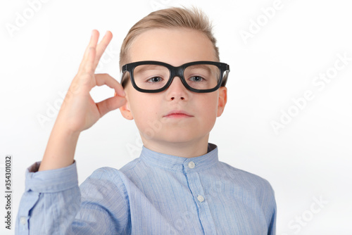 Cute caucasian boy with glasses in blue shirt shows gesture ok or zero.Photo studio white wall.