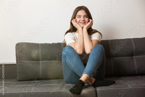 young beautiful smiling brunette woman sitting on the sofa in her appartment holding hands near face looking happy, feels cozy at home