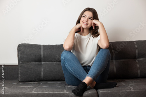 young beautiful smiling brunette woman sitting on the sofa in her appartment holding hands near face looking dreamy, feels cozy at home