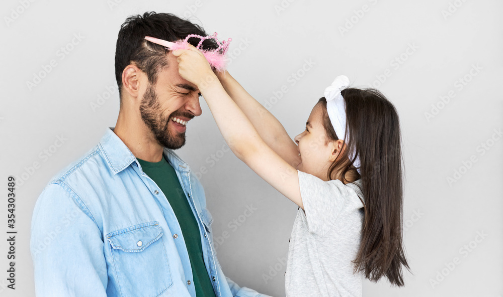 Obraz premium Portrait of lovely daughter smiling broadly puts the princess's crown on the head of her handsome father isolated over light gray background. Little girl and dad having fun on Father's Day.