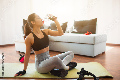 Young woman doing sport workout in room during quarantine. Rest after exercise. Girl sit on mat and drink water from plastic bottle. Pause after wokrout. photo