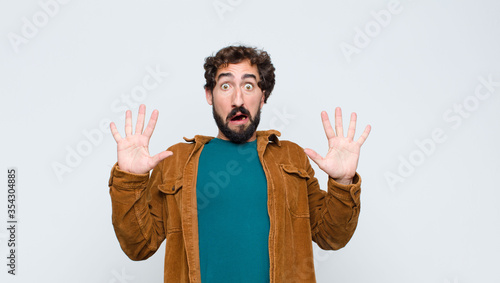 young handsome man feeling stupefied and scared, fearing something frightening, with hands open up front saying stay away against flat wall photo