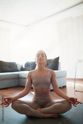 Young woman doing yoga workout in room during quarantine. Girl sit in lotus position and meditate with closed eyes. Stretching and exercising at home.
