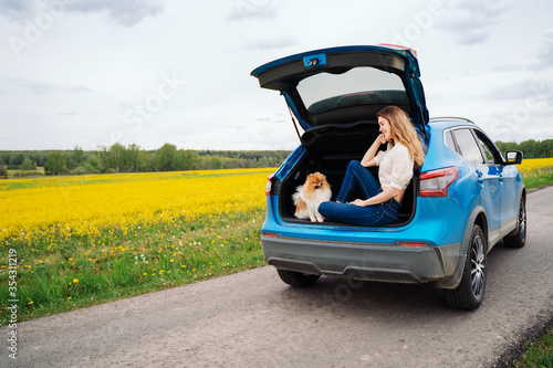 Happy young woman traveler with dog sits in car on spring road. Freedom and traveling couple. Cheerful woman in casual clothes travels with pets in summer break on car trips. Outdoor recreation