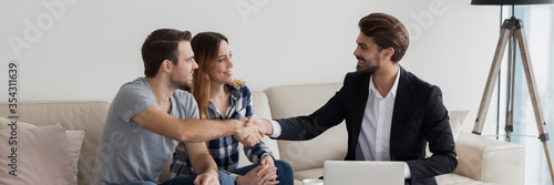 Happy couple young family flat renters shake hands with realtor seated on sofa in living room. Make real estate deal, first dwelling purchase concept. Horizontal photo banner for website header design