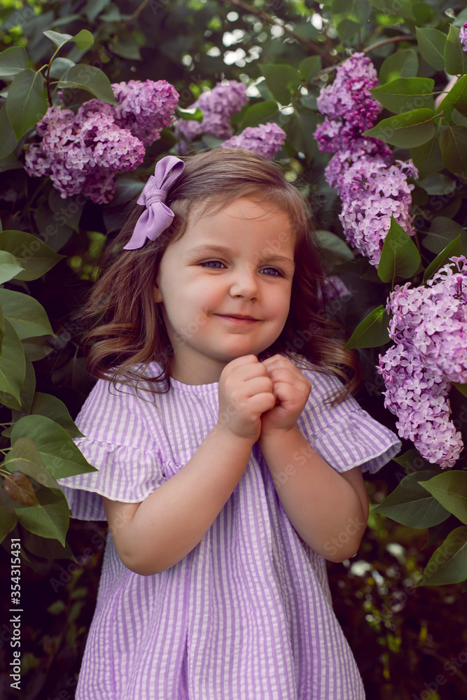 Fototapeta premium three-year-old girl stands in lilac bushes in a dress and a bow