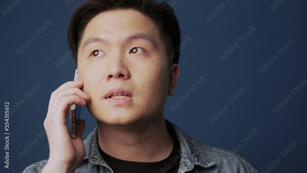 A close-up view of an attractive young asian man is talking on the phone isolated over dark blue wall background in studio A close-up view of an attractive young asian man is talking on the phone isolated over dark blue wall background in studio