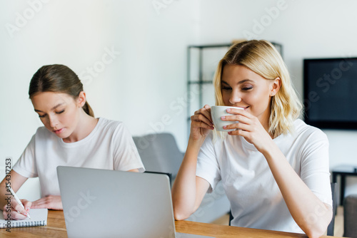 happy freelancer drinking coffee near sister writing in notebook