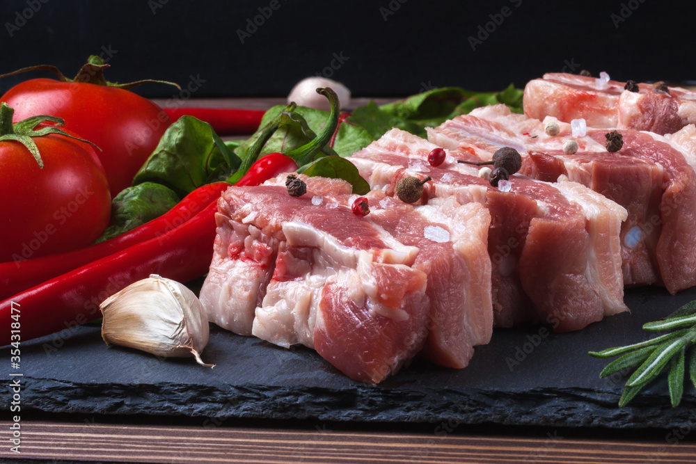 Raw slices of pork on a black slate board, tomatoes, red pepper and powder. Pork belly with vegetables.