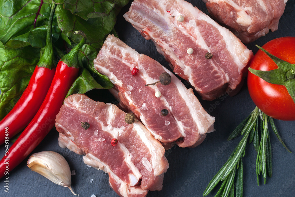 Raw slices of pork on a black slate board, green salad, tomatoes, rosemary, garlic, red pepper and powder. Pork belly with vegetables. Raw slices of pork on a black slate board, green salad, tomatoes, rosemary, garlic, red pepper and powder. Pork belly with vegetables.
