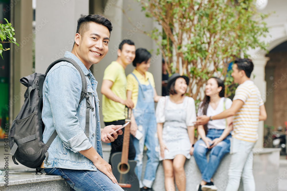 Obraz premium Portrait of handsome young Vietnamese university student with backpack texting and lookng at camera