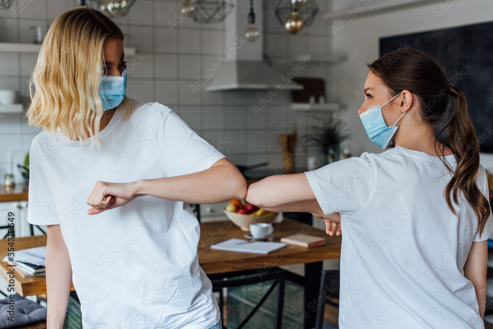 Fototapeta premium Sisters in medical masks greeting with elbow bump in living room