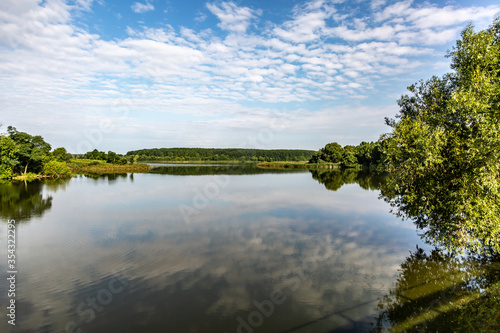 View of the river lake, green trees and bushes around and reflection of the sky with clouds on a sunny day. Horizontal orientation.