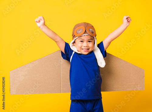 Happy Asian handsome funny child or kid little boy smile wear pilot hat play and goggles raise hand up with toy cardboard airplane wings flying, studio shot isolated yellow background, Startup freedom photo