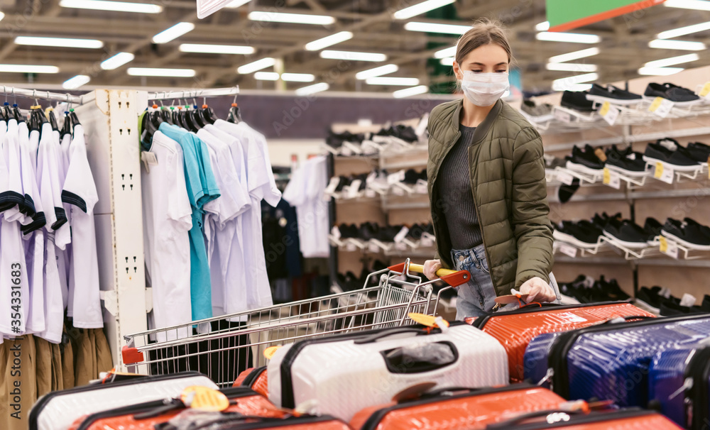 Social distancing concept. A young woman in a disposable mask choosing a suitcase for the upcoming vacation after the end of the Covid-19 coronavirus pandemic. Shopping at supermarket during epidemic
