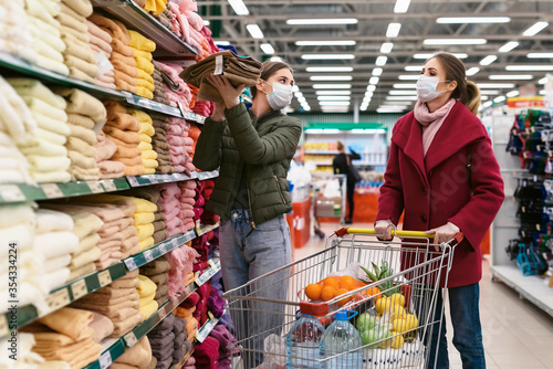 Social distancing in a supermarket. Young women in disposable face masks with grocery baskets choosing kitchen towels and household goods. Shopping during the Coronavirus Covid-19 epidemic