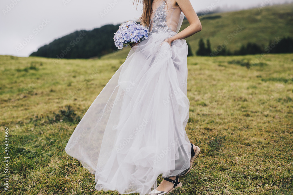 a young beautiful girl in a white dress looking like a bride walking through green meadows and hills with gartensia in her hands
